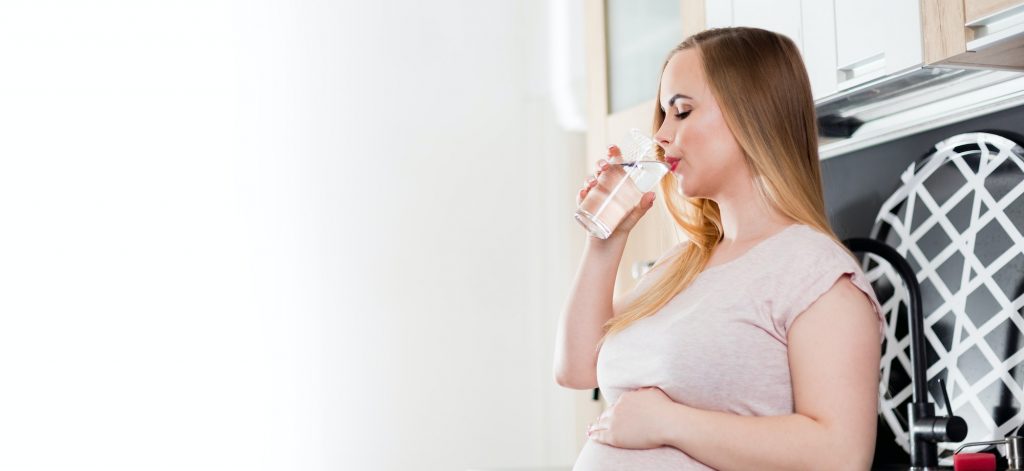Pregnant woman drinking fresh water in kitchen, healthcare during pregnancy concept